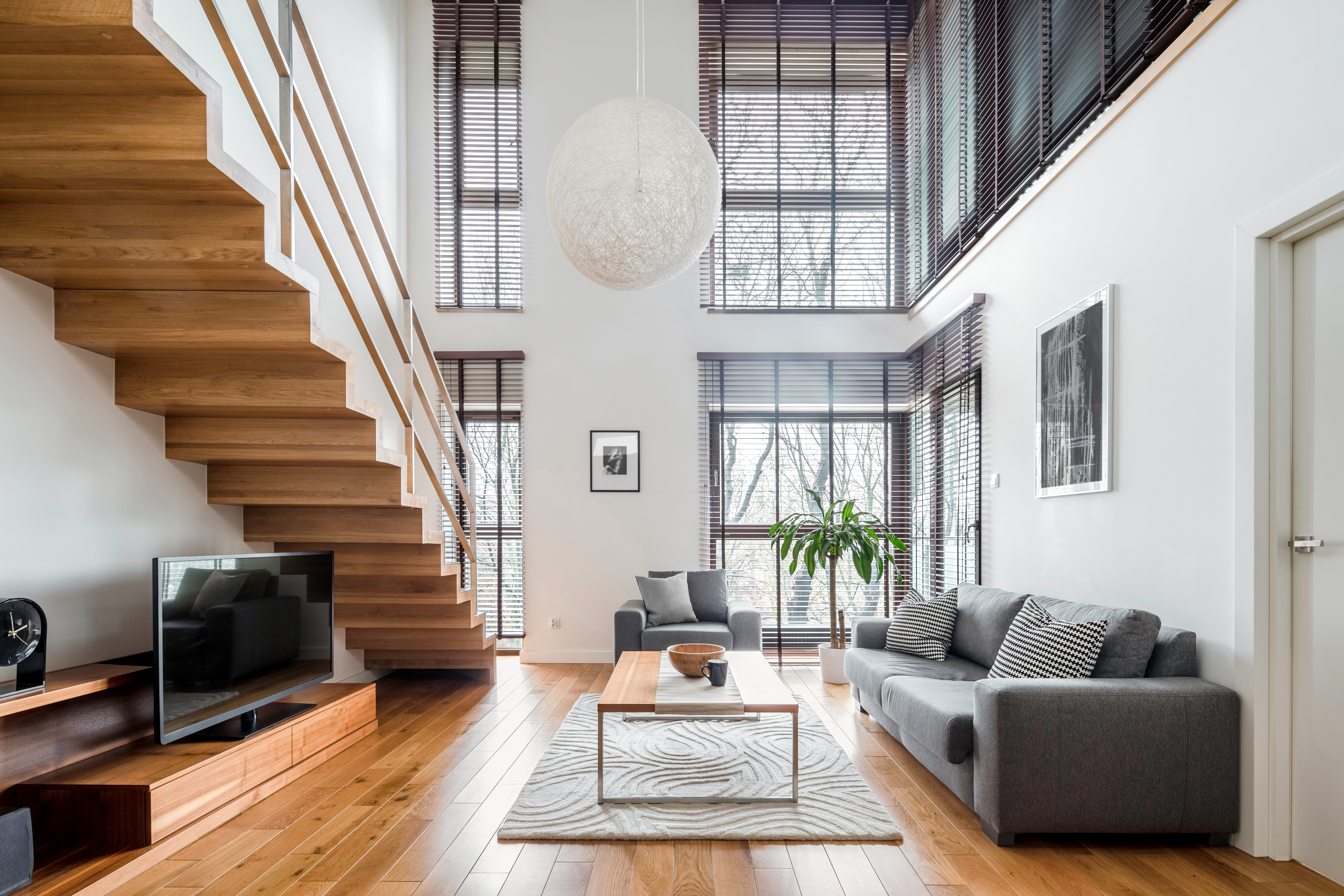 Modern loft living room with wooden stairs, gray sofa, and floor-to-ceiling windows with blinds